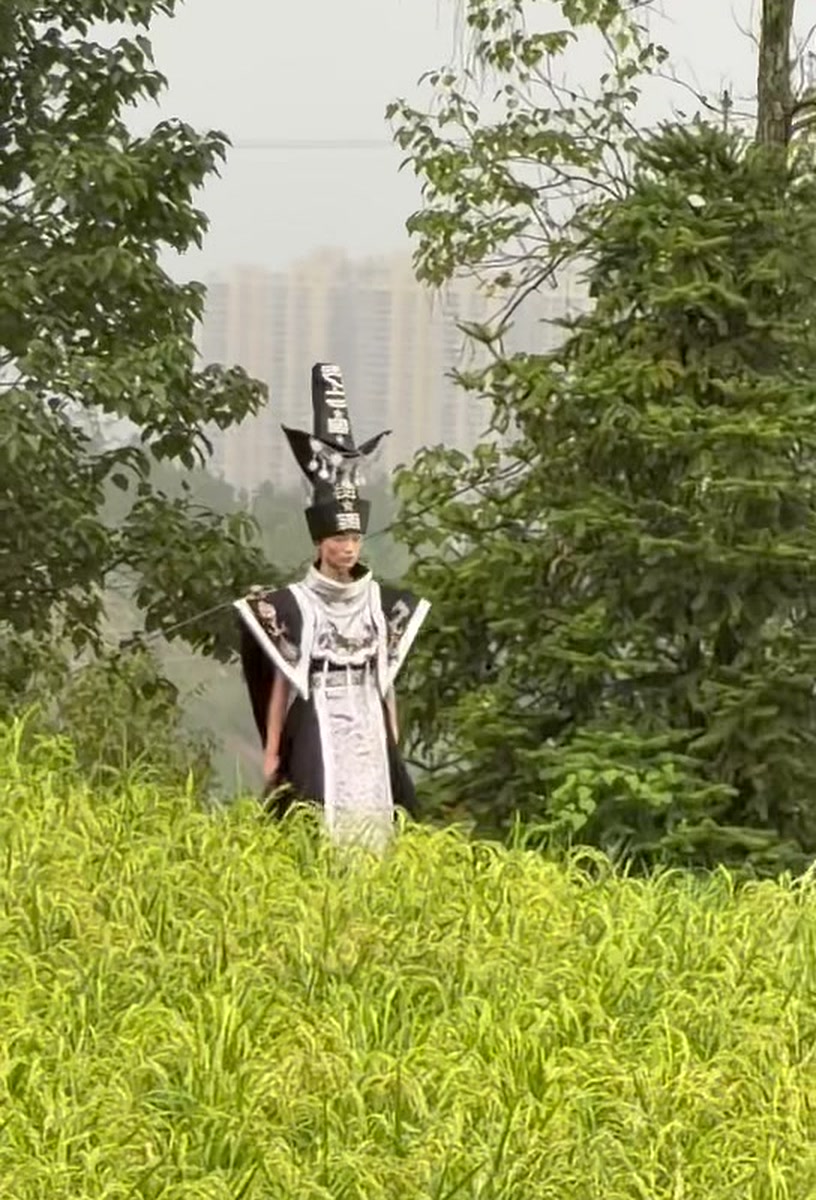 A model in an avant-garde Miao-inspired headdress, walking a runway laid through a rice paddy outside Guiyang, with the city skyline behind